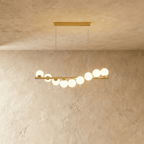 Clérachic gold linear chandelier with nine opal white glass globes on a brass bar, lit warm and suspended on two thin wires.