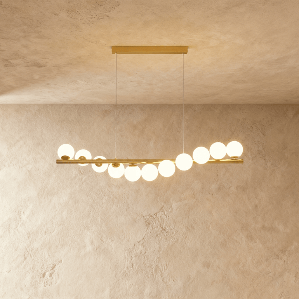 Clérachic gold linear chandelier carrying eleven opal white glass globes along a brass bar, lit warm over a limewash wall.