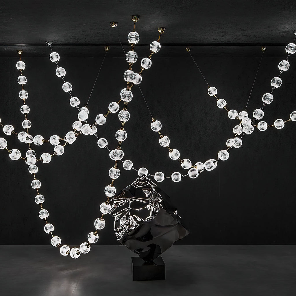 Against a black wall, loops of clear faceted spheres hang beside a dark sculpted form, the strands of this Clérachic pendant crossing at several heights.