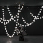 Against a black wall, loops of clear faceted spheres hang beside a dark sculpted form, the strands of this Clérachic pendant crossing at several heights.
