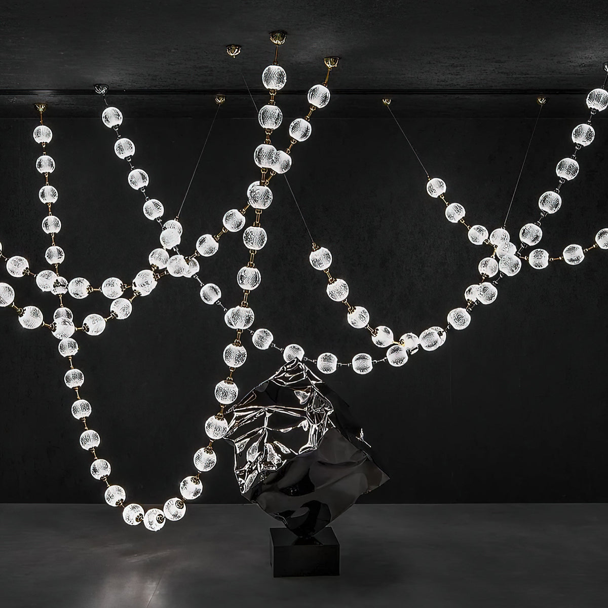 Against a black wall, loops of clear faceted spheres hang beside a dark sculpted form, the strands of this Clérachic pendant crossing at several heights.