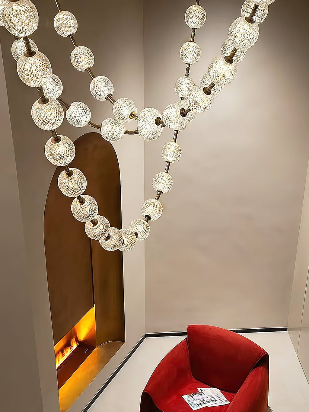 Textured spheres on gold links loop down past a tall arched niche with a glowing fire, a red armchair sitting below this Clérachic pendant.