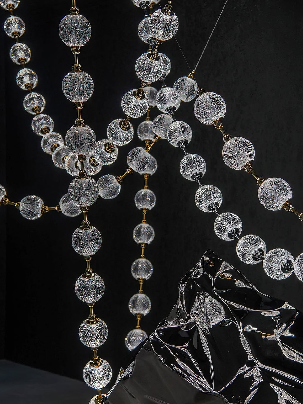 Close view of clear patterned spheres and gold connectors on crossing strands of this Clérachic pendant, with a dark reflective sculpture below.
