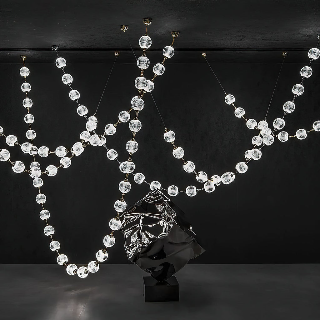 Wide view of this Clérachic pendant in a dark gallery space, its beaded strands swooping across the room above a mirrored sculpture on a black plinth.