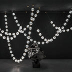 Wide view of this Clérachic pendant in a dark gallery space, its beaded strands swooping across the room above a mirrored sculpture on a black plinth.