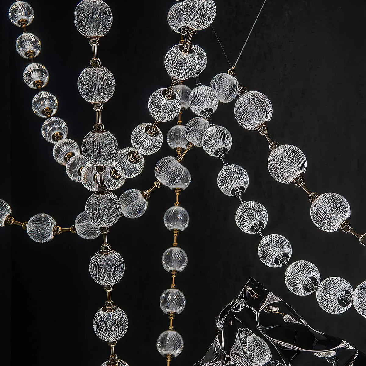Close view of the cut-pattern glass spheres and slim brass rods where several strands of this Clérachic pendant cross against a black wall.
