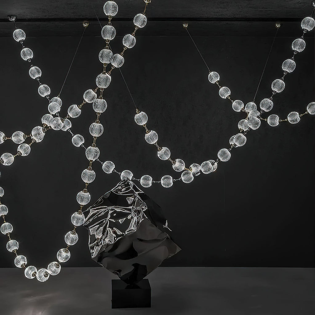 Lit strands of clear spheres sweep across a dark wall behind a crumpled metal sculpture, the full span of this Clérachic pendant on show.