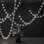Lit strands of clear spheres sweep across a dark wall behind a crumpled metal sculpture, the full span of this Clérachic pendant on show.