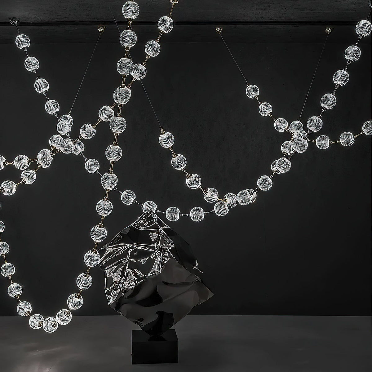 Lit strands of clear spheres sweep across a dark wall behind a crumpled metal sculpture, the full span of this Clérachic pendant on show.