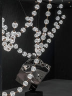 Looking up through crossing strands of this Clérachic pendant, patterned spheres glow in front of a black wall with a polished sculpture below.