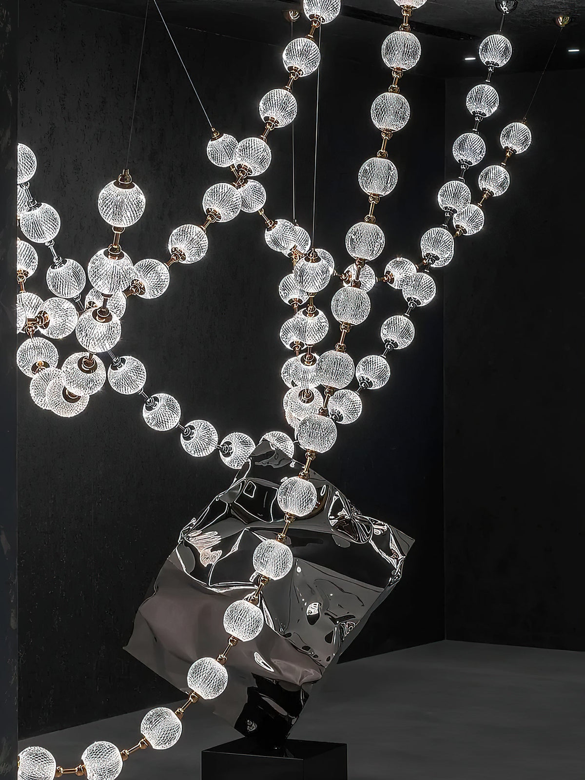 Looking up through crossing strands of this Clérachic pendant, patterned spheres glow in front of a black wall with a polished sculpture below.