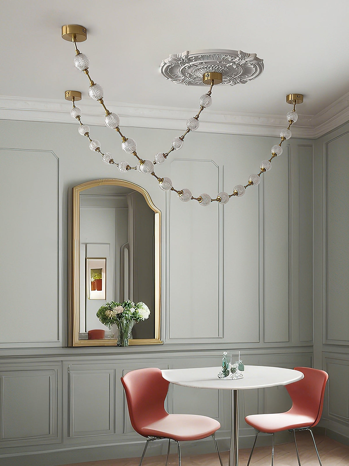 Two swags of frosted spheres hang below an ornate ceiling rose in a grey-panelled dining nook with a gold mirror and a white round table, from Clérachic.