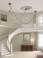 Beaded strands in frosted glass loop across a ceiling beside a curved white staircase and landing, a Clérachic pendant above a carved wooden sideboard.
