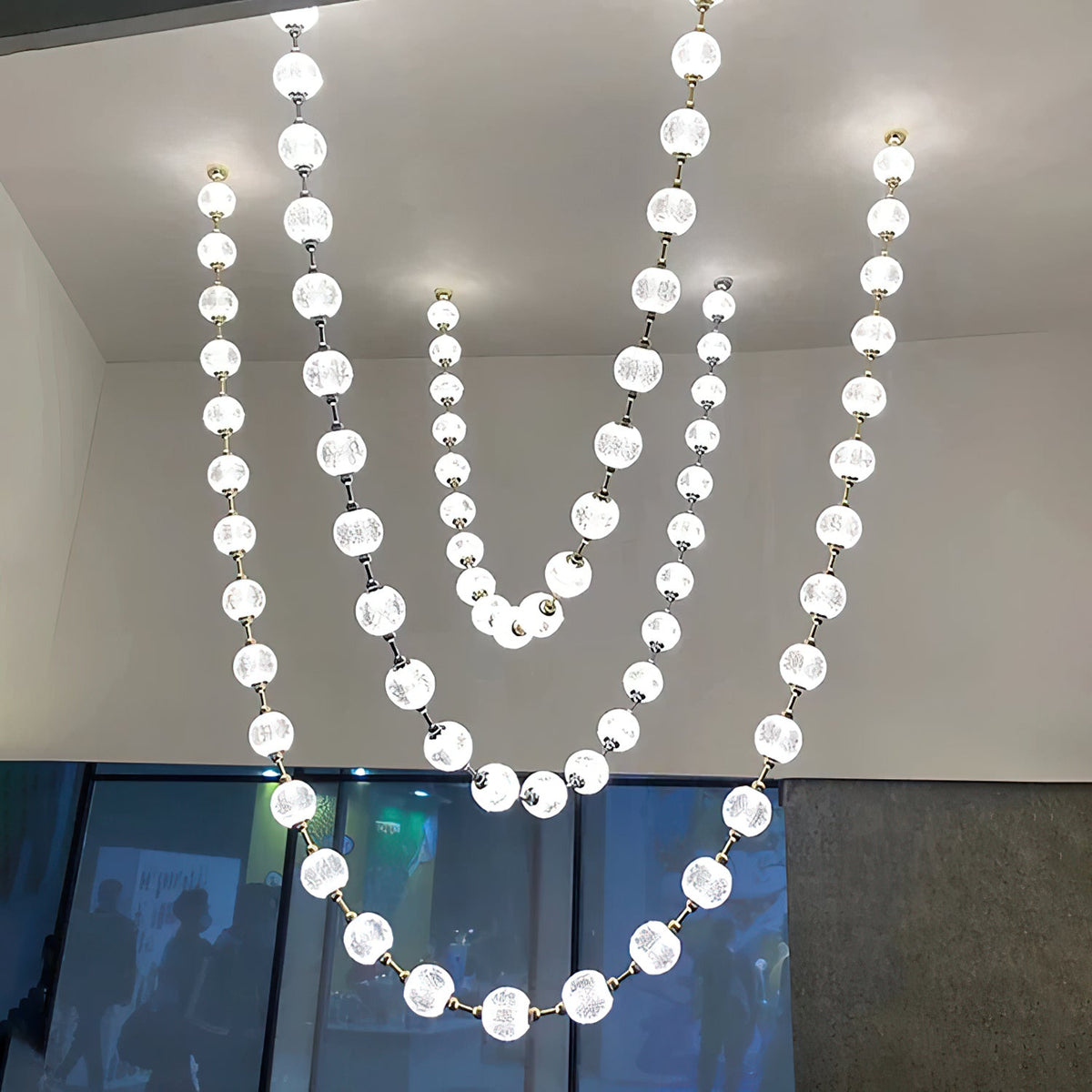 Looking up at four long, lit strands of white spheres from this Clérachic pendant, hung high in a large room with a window wall.