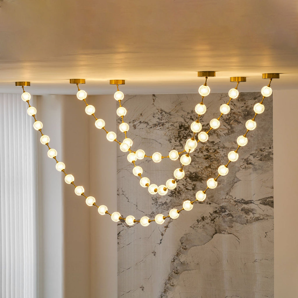 Warm-lit strands of spheres on gold links drop from a ceiling edge and drape in layers over a veined marble wall, from this Clérachic pendant.