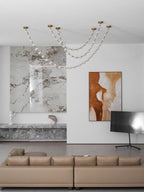Unlit strands of this Clérachic pendant sit high on the ceiling of a white living room with a marble feature wall and a tan sofa.
