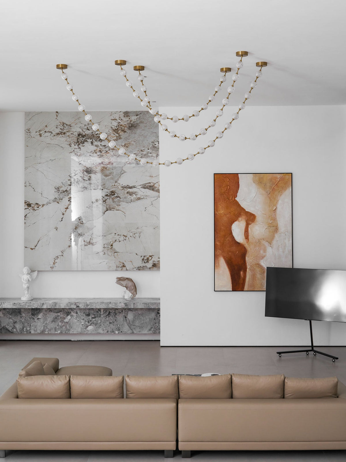 Unlit strands of this Clérachic pendant sit high on the ceiling of a white living room with a marble feature wall and a tan sofa.