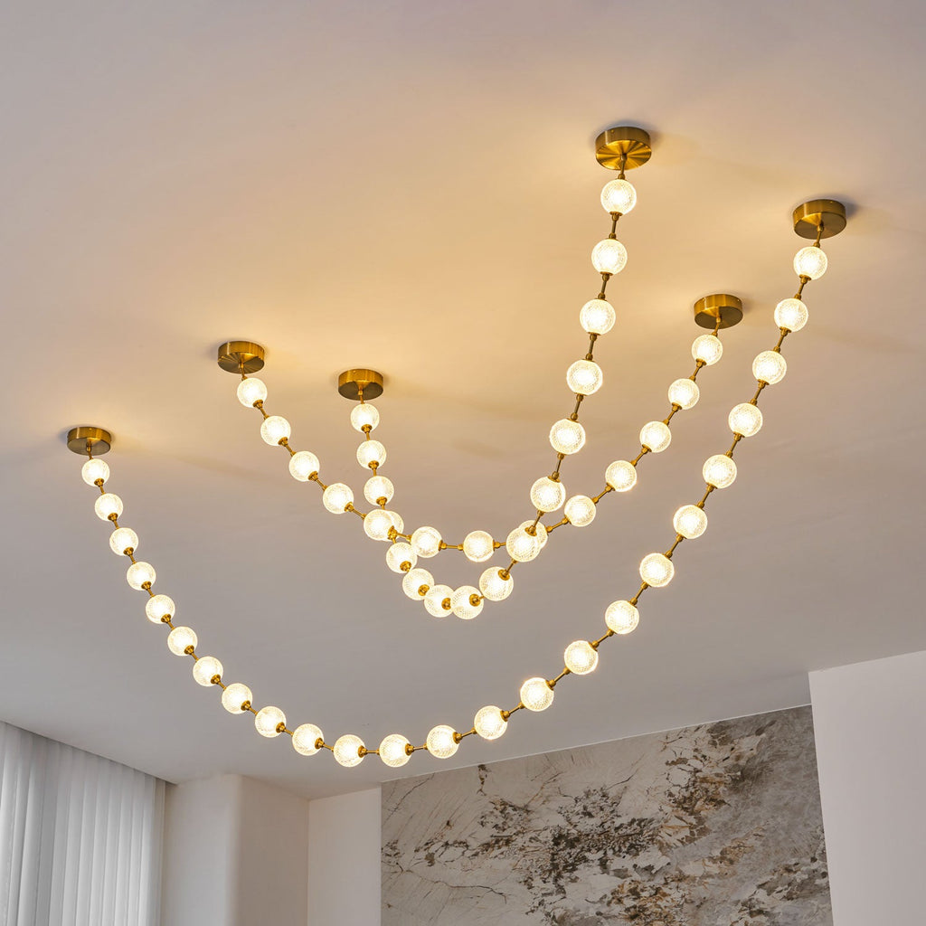 Lit warm white, this Clérachic pendant shows six strands of different lengths from separate brass plates, with a marble wall behind.