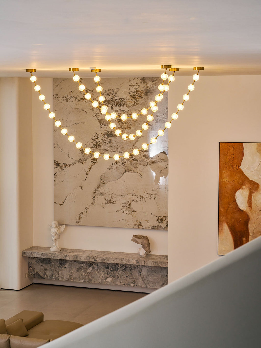 Warm-lit spheres on this Clérachic pendant loop down from the ceiling edge of a mezzanine, above a stone shelf, a small bust and a red-toned painting.