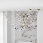 Unlit strands of frosted spheres fall across a veined marble wall beside sheer curtains, with this Clérachic pendant showing six brass ceiling plates.
