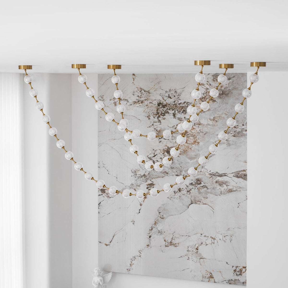 Unlit strands of frosted spheres fall across a veined marble wall beside sheer curtains, with this Clérachic pendant showing six brass ceiling plates.