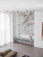 A wide room view shows the unlit strands of this Clérachic pendant against a marble wall, above a stone ledge and a low taupe sofa.