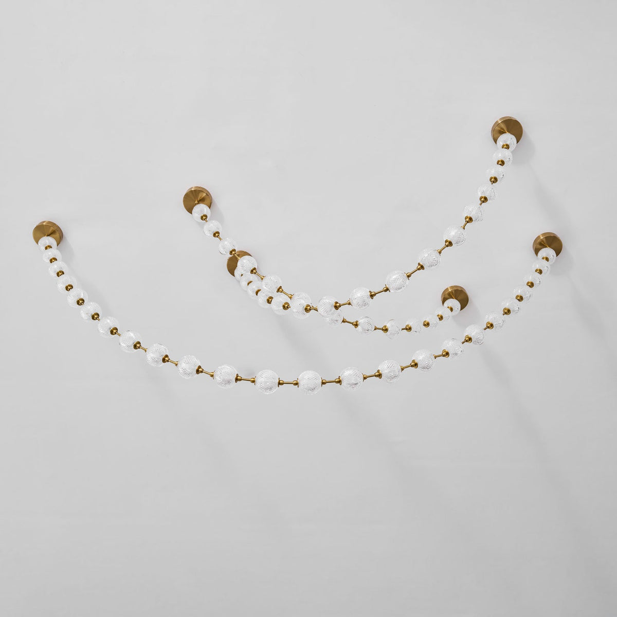 Plain white wall and ceiling corner with two unlit swags of frosted spheres on small brass plates, shown in a close crop of this Clérachic pendant.