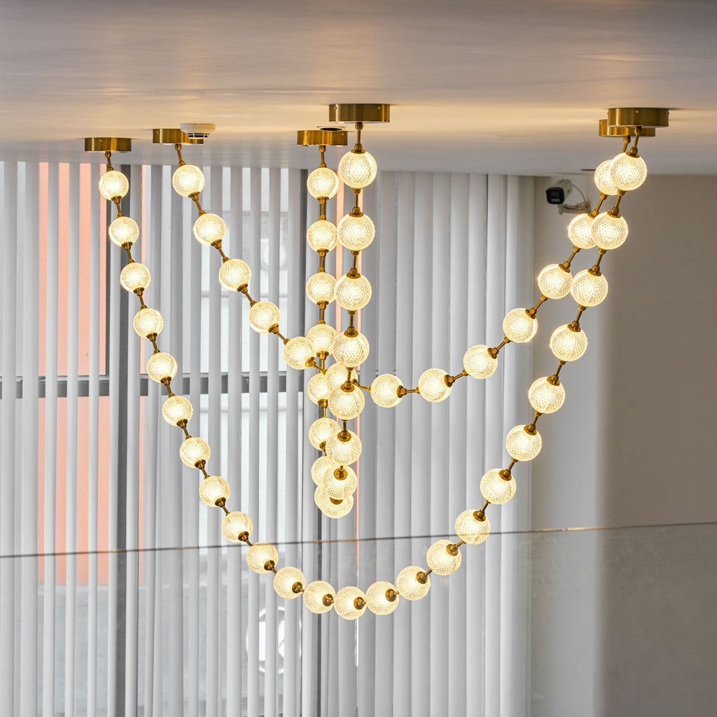 Glowing warm-white spheres in several overlapping loops hang in front of vertical blinds, a strong light from this Clérachic pendant.