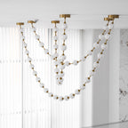 Unlit frosted spheres on gold rods hang in long loops in front of white vertical blinds, one of the swags on this Clérachic pendant falling low.