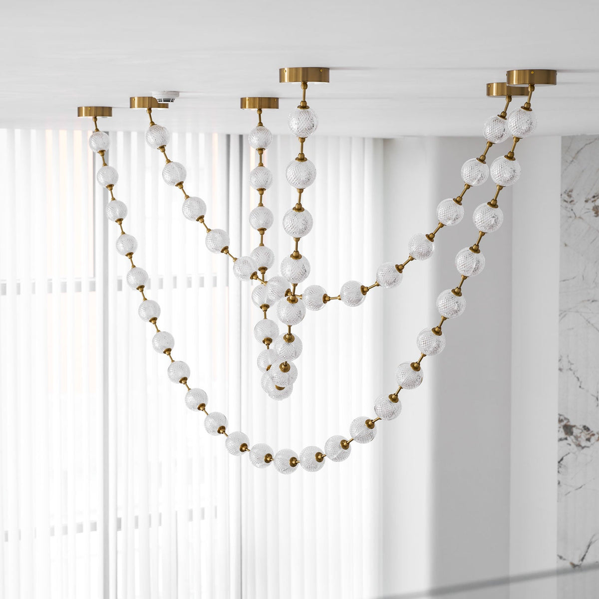 Unlit frosted spheres on gold rods hang in long loops in front of white vertical blinds, one of the swags on this Clérachic pendant falling low.