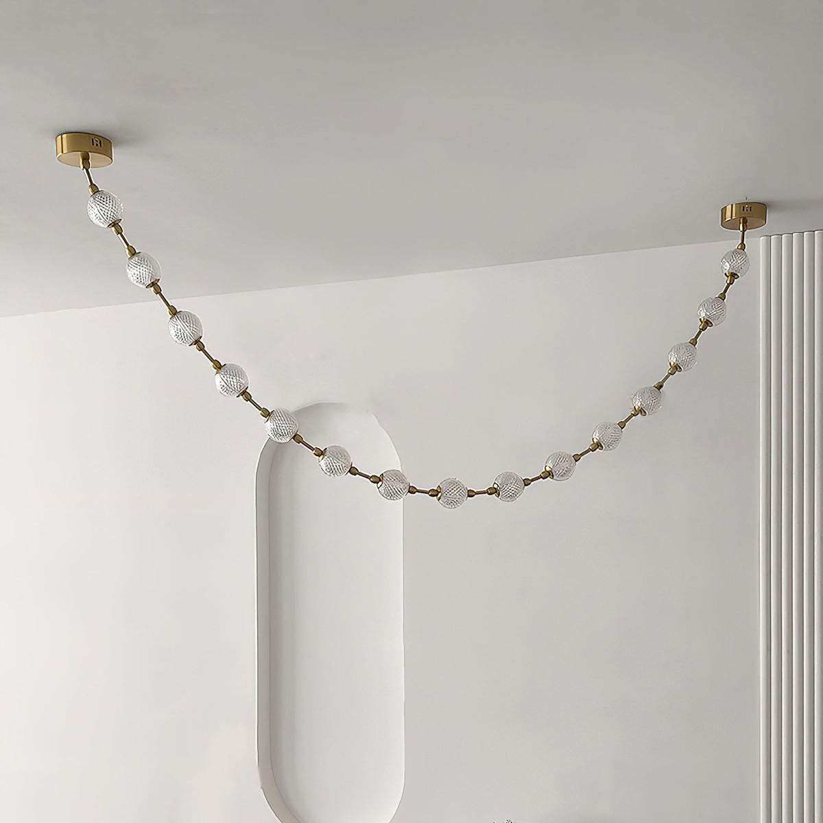 Looking up, a single swag of frosted spheres on gold rods spans a white ceiling between two brass plates, from this Clérachic pendant.
