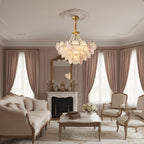 A Clérachic pendant with pink and frosted discs hangs in a living room with a cream sofa, pink curtains and a marble fireplace.