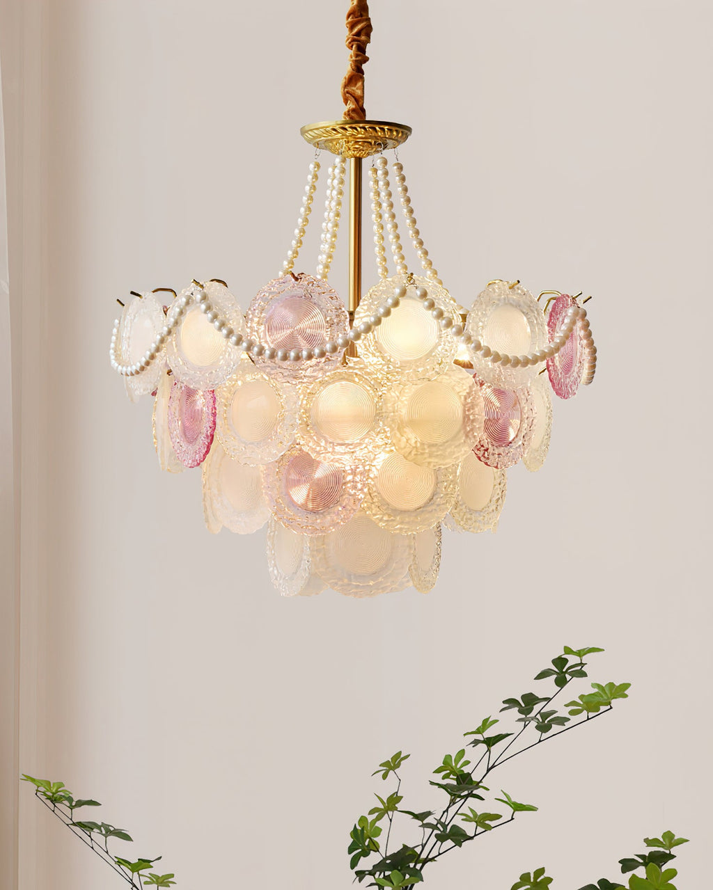 Clérachic pendant with a gold canopy, pearl strands and tiered glass discs, hung against a plain wall with green leaves below.