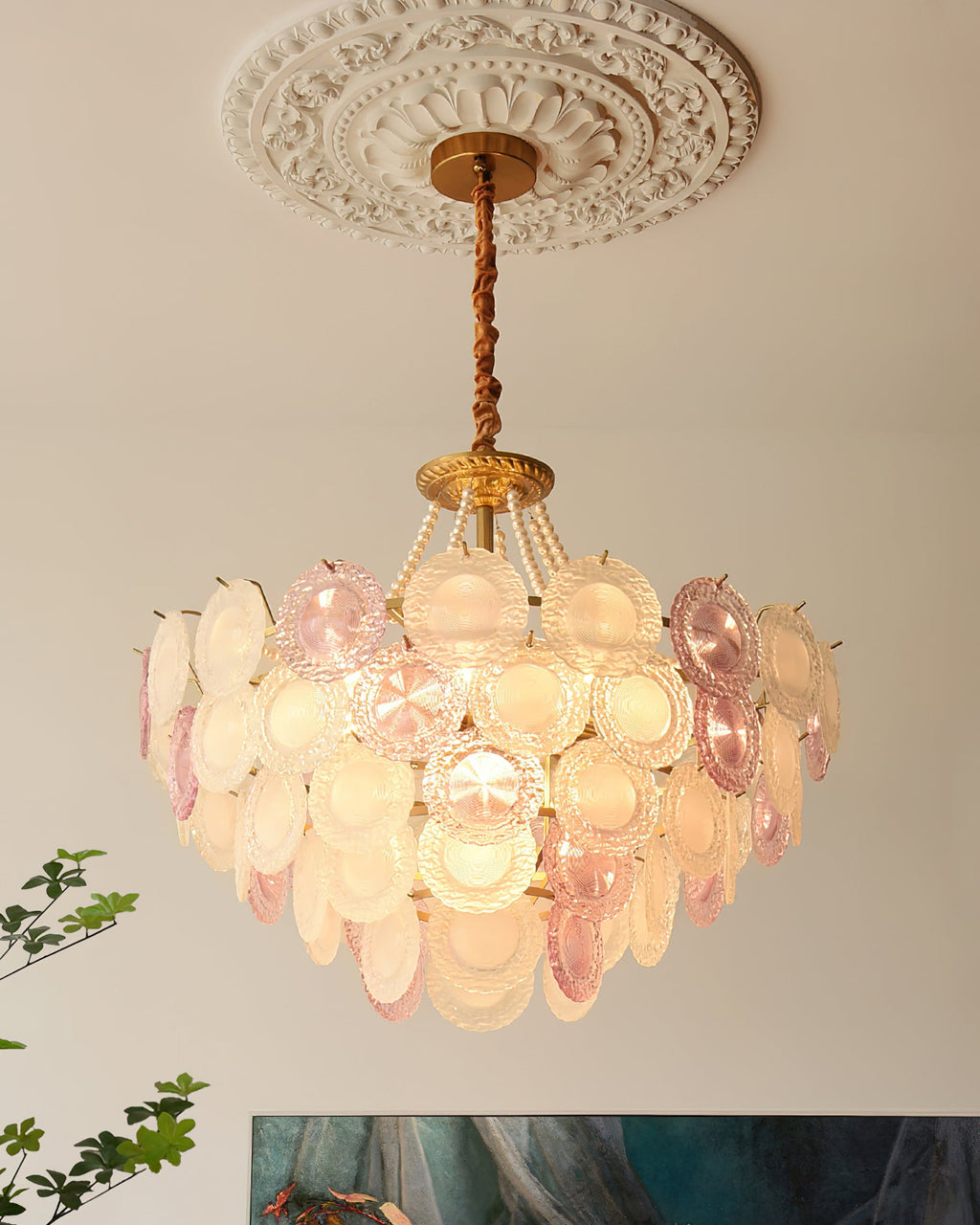 This Clérachic pendant hangs below an ornate plaster ceiling rose, its glass discs glowing warm, with a blue-green painting at the edge.
