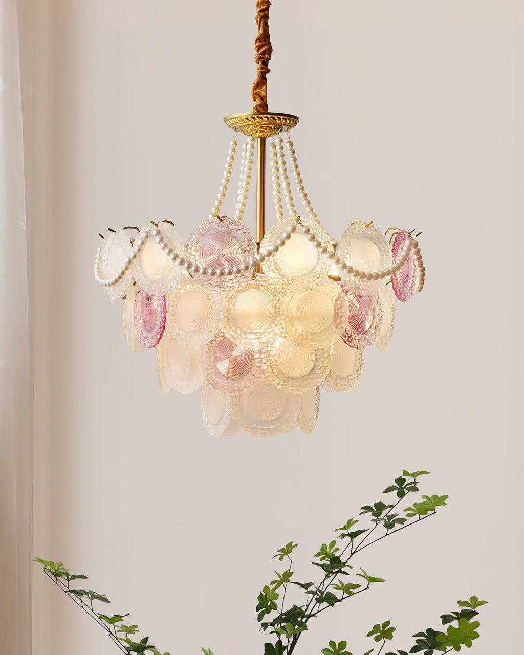 Lit Clérachic pendant against a pale wall with a soft curtain, its glass discs glowing and a leafy branch rising below.