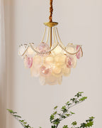 Lit Clérachic pendant against a pale wall with a soft curtain, its glass discs glowing and a leafy branch rising below.