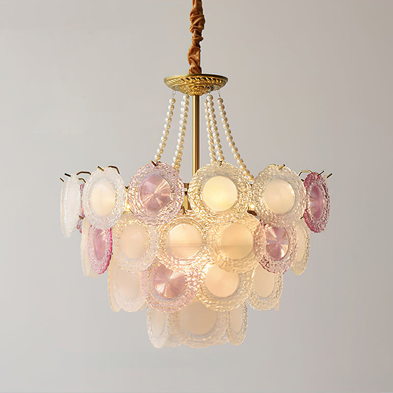A lit Clérachic pendant with a gold canopy, pearl strands and layered pink and frosted glass discs, on a pale grey backdrop.
