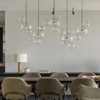 Several Clérachic glass sphere pendants with gold stems hung over a long dining table with olive-green armchairs.
