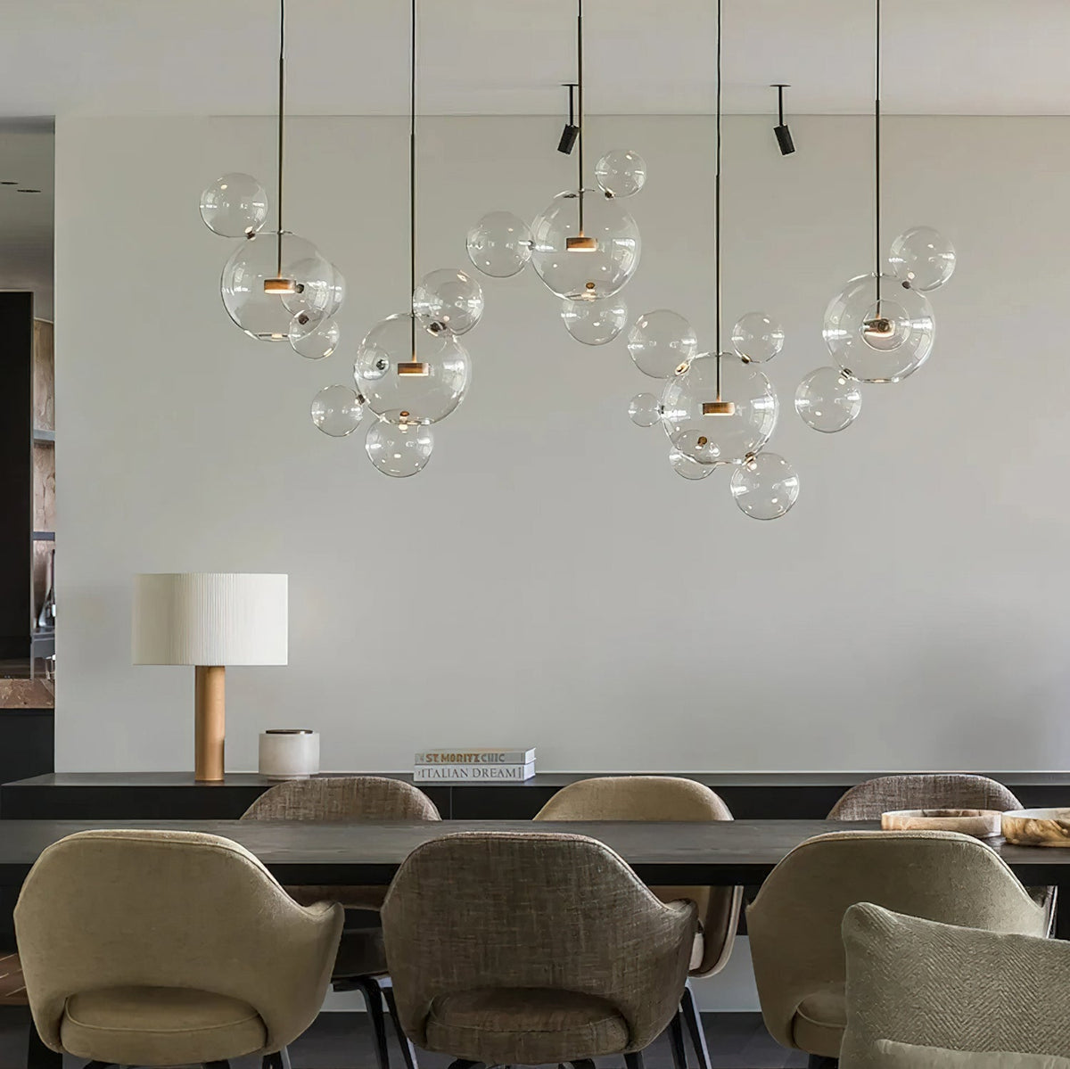 Several Clérachic glass sphere pendants with gold stems hung over a long dining table with olive-green armchairs.