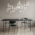 Wide cluster of Clérachic glass sphere pendants above a black dining table, with black chairs on a pale wooden floor.