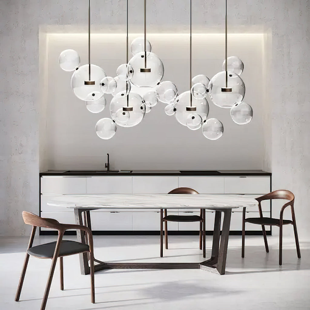Clérachic glass sphere pendants over a marble-topped dining table with wooden chairs, under a lit recess in a pale wall.