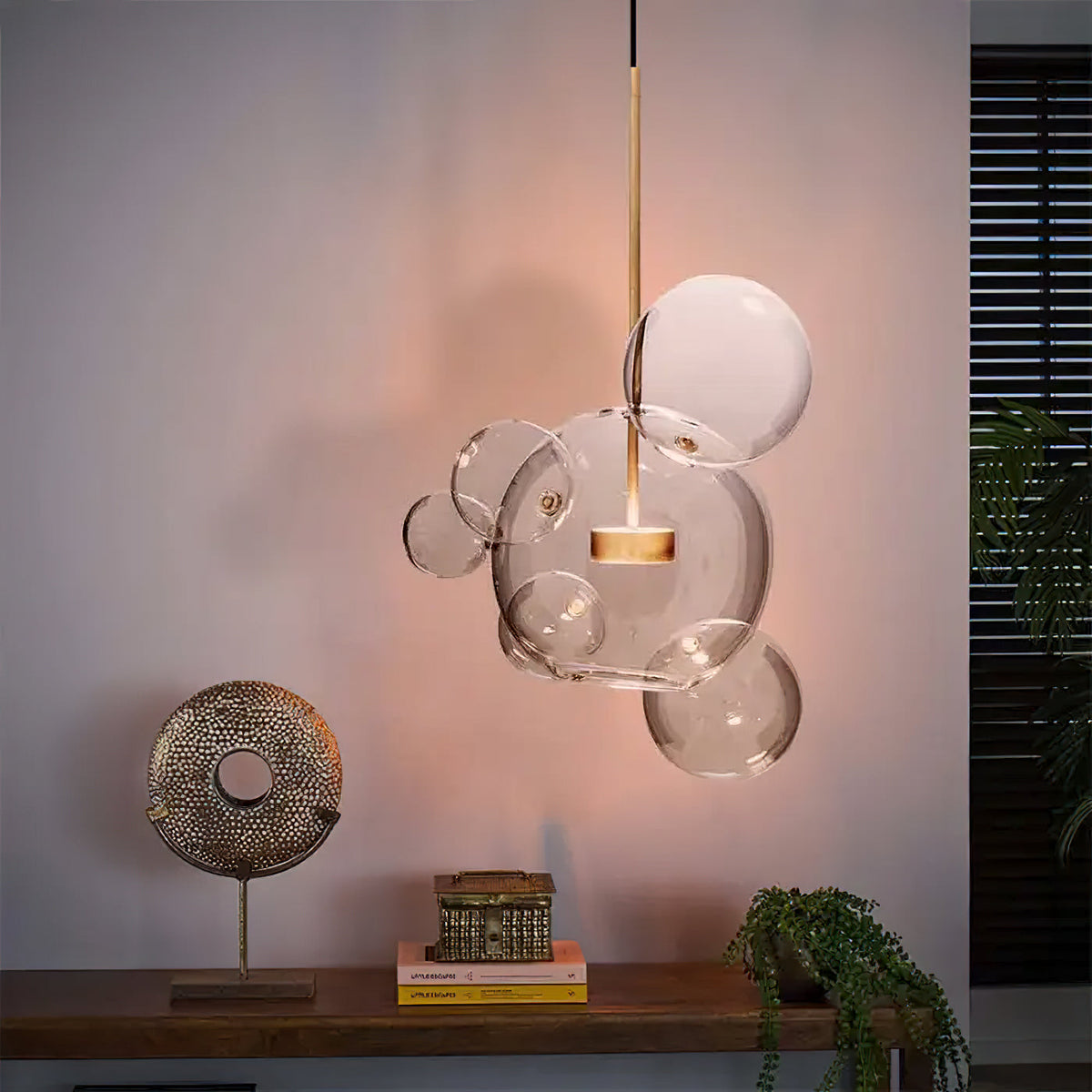 Lit Clérachic glass sphere pendant against a warm pink wall, above a wooden shelf with a brass disc sculpture and a plant.