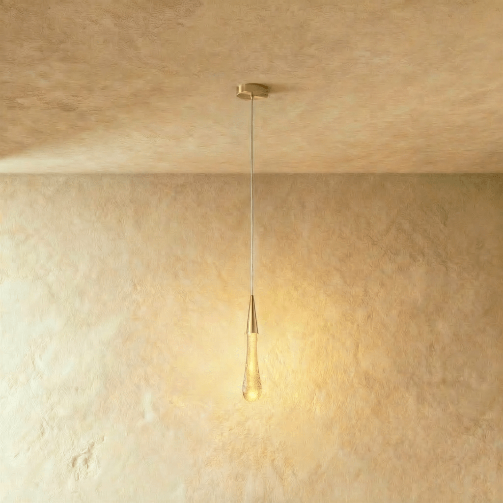 Clérachic gold teardrop pendant with a bubbled glass drop and round brass canopy, glowing warmly on a limewash wall.
