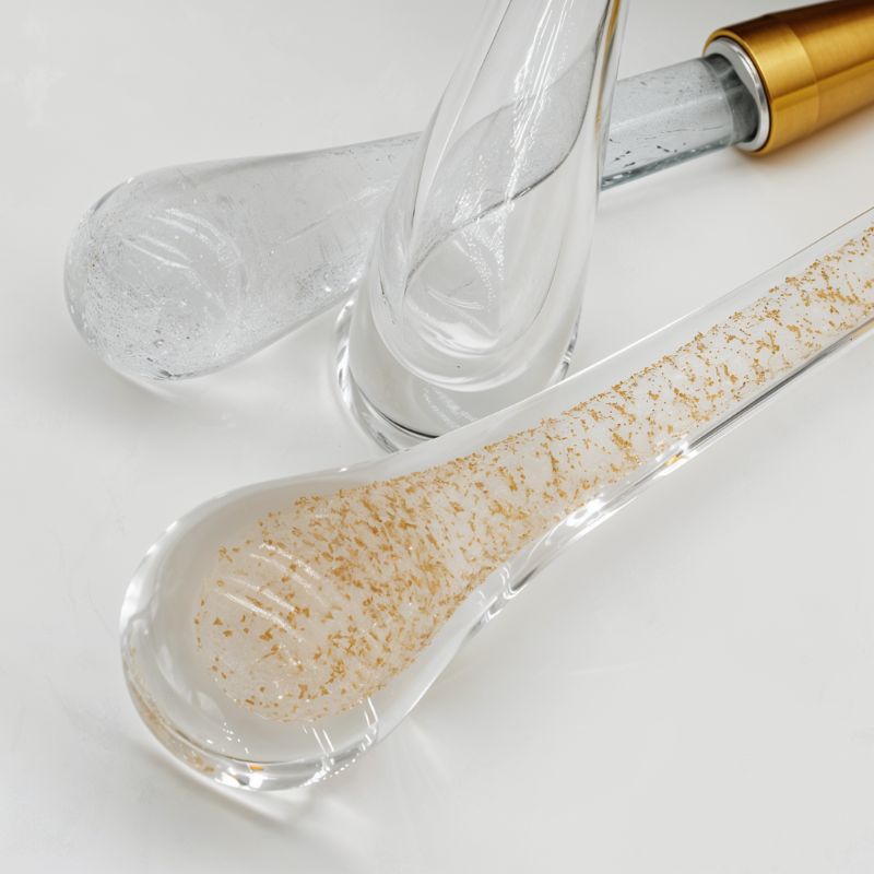 Clérachic close-up of clear teardrop glass drops, one flecked with gold leaf, beside a brass cap on a white surface.