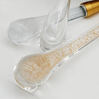Clérachic close-up of clear teardrop glass drops, one flecked with gold leaf, beside a brass cap on a white surface.