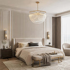 A wide bowl-shaped glass-rod pendant hangs in a calm bedroom with a ribbed headboard wall, a cream bed and a gold-framed bench, from Clérachic.