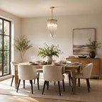 A slim Clérachic pendant with looped glass rods hangs above a wooden dining table with cream chairs, in a room with a black-framed glazed door.