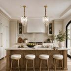 A pair of narrow Clérachic pendants hang above a marble kitchen island with white stools and brass-footed legs, in a cream kitchen.