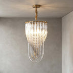 A narrow Clérachic pendant with glass rods looping to a point and a twisted gold stem hangs in a grey plaster room, lit warm.