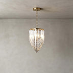 The smallest Clérachic pendant, with a slim glass-rod body ending in a point, hangs on a fine gold chain against a grey plaster wall.